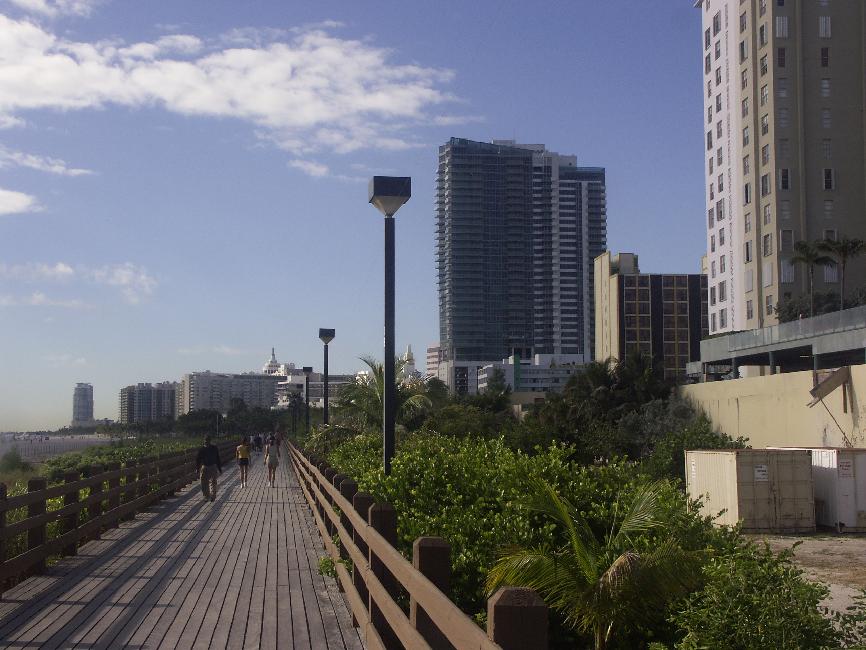 Promenade entre les immeubles et les piscines à droite, une bande de terre et la plage à gauche