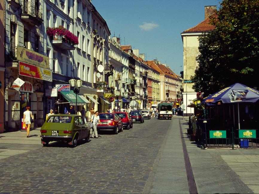 La rue commerçante de Kalisz ; compte tenu de la température encore estivale, la place qui se trouve à droite est couverte des terrasses des bistros.Les voitures sont les mêmes que chez nous, avec une densité plus forte de vieilles Fiat 500. Par contre, on croise sur les routes de nombreux camions antiques, poussifs et puants.